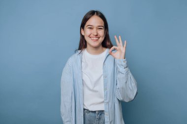 Genç bir kadın gülümsüyor ve sıradan bir kıyafetle mavi bir arka plana karşı iyi bir jest yapıyor.