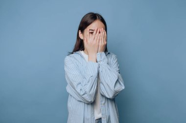 Genç bir kadın, evdeki sıradan bir an sırasında mavi bir arkaplan önünde duygularını ifade ederek ellerini yüzünü kapatıyor.