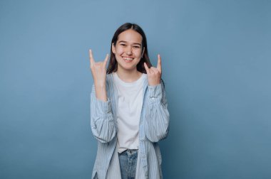 Genç bir kadın mavi arka planda gülümserken taş el hareketi yapıyor.