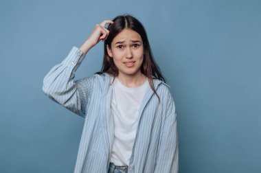 Kafası karışmış genç bir kadın, mavi bir arkaplanda dikilirken şaşkın bir ifade takınır.