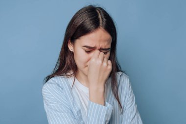 Bir kadın gözlerini kapalı tutarak mavi bir arka plan önünde sıkıntısını ifade eder.