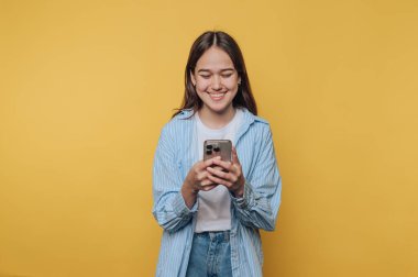 Çizgili tişört giyen gülümseyen genç bir kadın, parlak sarı bir fon önünde akıllı telefonuyla etkileşime girer.
