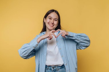 Gülümseyen genç bir kadın, ellerini parlak sarı bir arka plana koyup kalp şeklinde bir kalp yaparken gündelik tarzını gösteriyor.