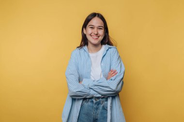 Mavi çizgili gömleğiyle, parlak sarı bir arkaplanda kolları bağlı duran genç bir kadın, kendinden emin bir şekilde gülümsüyor.