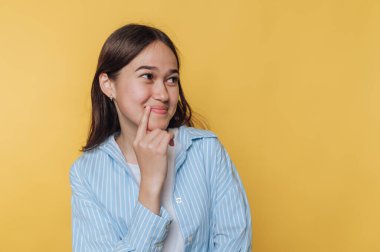 Neşeli ifadeli genç bir kadın parlak sarı arka plana karşı düşüncelerini ve fikirlerini düşünürken poz veriyor.