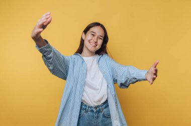 Açık kollarla gülümseyen genç bir kadın parlak sarı bir arka plana karşı neşeli bir iç mekan ortamında