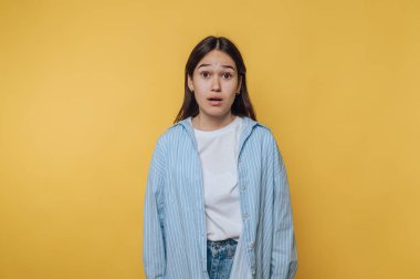 Kapalı alanda çekilen bir fotoğraf çekiminde parlak sarı bir arkaplan önünde şaşırdığını ifade eden genç bir kadın.