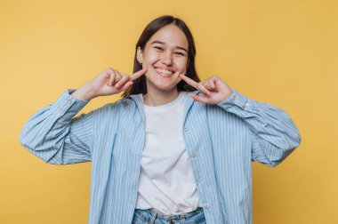 Genç bir kadın gülümser ve parlak sarı bir arka plana karşı poz verir. Çizgili gömlek ve günlük kot pantolon giyer.