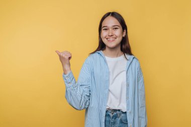 Gündelik elbiseli genç kadın gülümseyip, parlak sarı bir arka plana işaret ediyor.