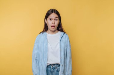 Şaşırmış genç bir kadın, kapalı bir stüdyoda çekilen bir fotoğraf çekiminde parlak sarı bir arkaplan önünde şaşkınlığını dile getiriyor.