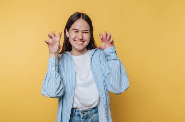 Komik bir şekilde gülümseyen kadın ev içi fotoğraf çekimi sırasında sarı arka planda el pençe yapıyor.