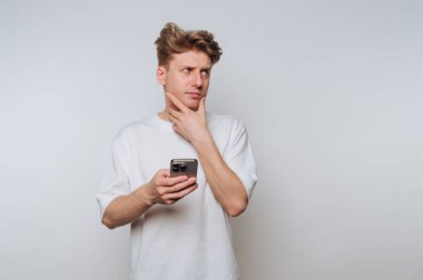 Genç adam minimalist bir stüdyo ortamında akıllı telefon tutarken düşünüyor.