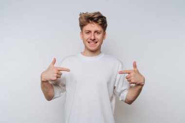 Genç adam düz beyaz bir arka plana karşı duruyor, iki eliyle kendini göstererek geniş bir gülümseme içinde..