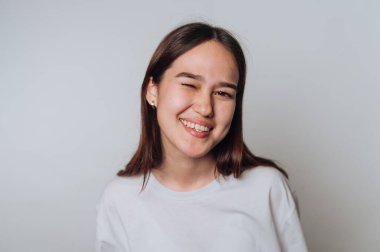 Gülümseyen genç bir kadın, arka planda hafif bir poz verirken kameraya göz kırpıyor.