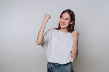 Kadın başarıyı neşeli bir ifadeyle kutluyor ve tarafsız bir arkaplanda yumruklarını kaldırıyor.