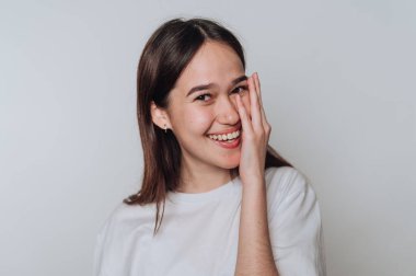 Genç bir kadın gülümsüyor ve bir eliyle yüzünü kapalı bir mekanda neşeyle örtüyor.