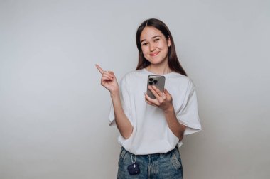 Genç bir kız gülümseyerek el kol hareketi yapıyor. Elinde akıllı bir telefon var ve evde takılıyor.