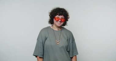 Turuncu güneş gözlüğü takan ve gri gömlekli neşeli genç bir kadın beyaz arka planda pozitiflik yayarak geniş bir gülümseme sergiliyor.