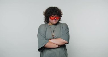 Gri tişörtlü genç kıvırcık kadın kollarını çapraz yapıyor, kendine güveni ve beyaz arka plandaki cesur ifadesiyle güçlü bir varlık sergiliyor.