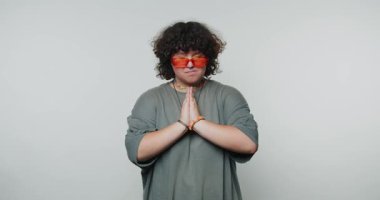 Düşünceli genç kadın ellerini dua etmek için bir araya getiriyor. Beyaz arka planda şık güneş gözlüğü takarken sakin ve düşünceli bir ruh hali sergiliyor.