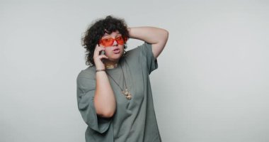 Genç kıvırcık kadın telefonda konuşuyor. Canlı turuncu güneş gözlüğü ve beyaz arka planda büyük beden bir gömlek takıyor.