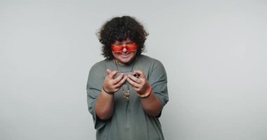 Genç kıvırcık kadın elinde akıllı bir telefonla neşeli bir yüz ifadesi sergiliyor. Beyaz arka planda bir anlık mutluluğu kutluyor.