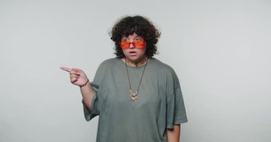 Genç kıvırcık kadın çarpıcı bir şekilde dışa bakıyor, büyük güneş gözlüğü takıyor, beyaz arka planda yön duygusunu aktarıyor.