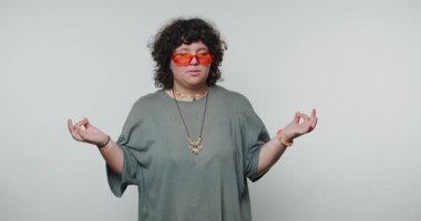 Kolları açık, elleri açık, turuncu güneş gözlüğü takan genç kıvırcık kadın, beyaz arka planda sakinlik ve farkındalık hissi yayıyor.