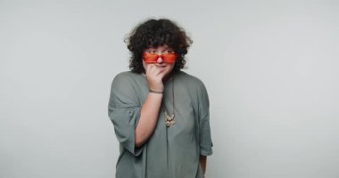 Kırmızı güneş gözlüğü takan kıvırcık saçlı genç bir kadın beyaz arka planda neşeli ve kaygısız bir ifade sergiliyor.