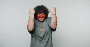 Neşeli genç kıvırcık kadın yumruklarıyla kutlama yapıyor, beyaz arka planda kırmızı güneş gözlüğü takıyor.