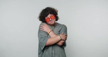 Kırmızı güneş gözlüklü genç kıvırcık kadın sarılıyor, beyaz arka planda kendini sevme ve rahatlama gösterisi yapıyor.