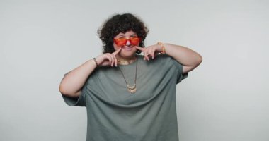 Genç kıvırcık kadın, beyaz arka planda neşe ve yaratıcılığı yansıtan neşeli bir parmak hareketi sergiliyor.