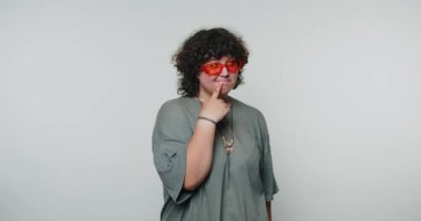 Yaramaz ama düşünceli bir ifade, beyaz arka planda kırmızı güneş gözlüğü takan genç kıvırcık kadın.