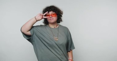 Kıvırcık saçlı ve güneş gözlüklü genç kıvırcık kadın parmaklarıyla şakacı hareketler yapıyor. Yaratıcılığını ve kaprislerini beyaz arka planda somutlaştırıyor.