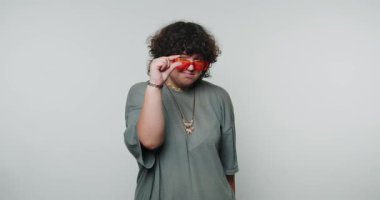 Genç kıvırcık kadın güneş gözlüklerini düzeltirken neşe içinde arkasına bakar, beyaz arka planda özgüven ve cazibenin bir karışımını yansıtır.