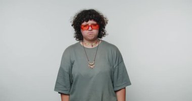 Neon güneş gözlüklü genç kıvırcık kadın ciddi bir ifade sergiliyor, beyaz arka planda derin bir tefekkür yansıtan ilgi çekici bir ruh hali yakalıyor.