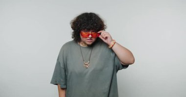 Genç kıvırcık kadın bir merak jesti sergiliyor, neon güneş gözlükleriyle süslenmiş, eşsiz duygusal perspektifleri vurguluyor ve beyaz arka planda yer alıyor.