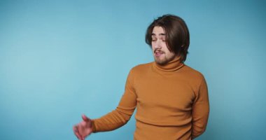 Turuncu kazaklı genç adam kendine güvenerek gülümsüyor, mavi arka planda açıklık ve çekiciliği öneriyor.