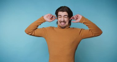 Turuncu balıkçı yakalı genç adam mavi arka planda kulaklarını sıkıntılı bir ifadeyle kapatarak duygularıyla boğuşuyor.