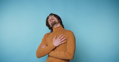 Genç adam kendine güvenen bir balıkçı yaka giyiyor, mavi arka planda dururken rahat bir yüz ifadesi sergiliyor.