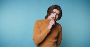 Düşünceli genç adam mavi arka plana bakarken çenesini sıvazlıyor.