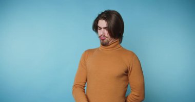 Balıkçı yaka kazaklı genç adam pazılarını esnetiyor ve mavi arka planda kendine güvenini gösteriyor.