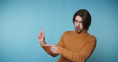 Kahverengi balıkçı yakalı genç adam şakacı bir şekilde işaret ediyor. Mavi arka planda hareket ve keyif olduğunu gösteriyor.