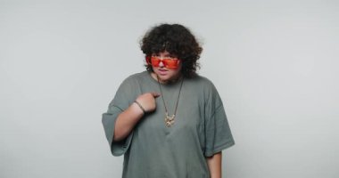 Kıvırcık şişman kız kendini işaret ediyor ve beyaz arka plandaki kameraya hoşnutsuzca bakıyor. Modern bir iç mekanda şık turuncu güneş gözlüğü takan kişi kendinden emin bir şekilde işaret eder.