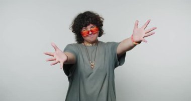 Gözlüklü komik kıvırcık kadın sarılmak istiyor, kameraya elini uzatıyor ve gülümsüyor. Güneş gözlüklü gülümseyen kadın stüdyoda yaratıcı bir etkinlik sırasında neşeli bir dansı paylaşıyor.
