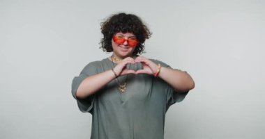 Genç bir kadın, kapalı bir alanda şık güneş gözlüğü takarak kalbi elleriyle şekillendirir.