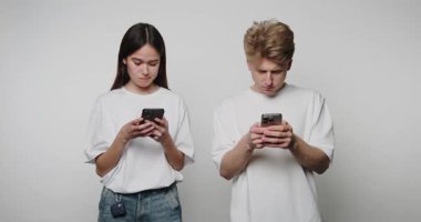 İki genç yetişkin yan yana duruyor, ikisi de akıllı telefonlarıyla meşgul. Gündelik beyaz tişörtler giyiyorlar ve modern iletişimi sergileyerek cihazlarının içine çekilmiş gibi görünüyorlar..