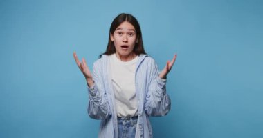 Etkileyici bir kadın, canlı bir ortamda mavi bir arkaplan karşısında heyecanlı bir şekilde el kol hareketi yapar. Çekici Asyalı kızın mavi arka planda kafası karışıyor ve anlamayan bir yüzü var ve kollarını yanlara açıyor.
