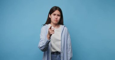 Çekici Asyalı kızın mavi arka planda kafası karışmış ve canı sıkkın bir şekilde kameraya parmağını gösteriyor.