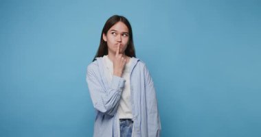 Genç bir kadın mavi bir arka plana karşı düşünceli bir yüz ifadesiyle poz veriyor ve sıradan bir kıyafet giyip kameraya bakıyor.